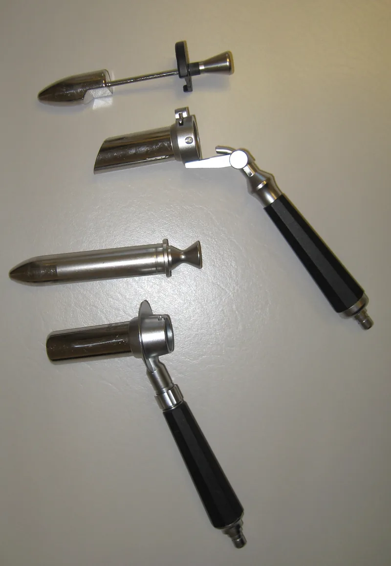 Two proctoscopes, short metal tubes with handles, each beside the smooth, rounded insert that fills the tube